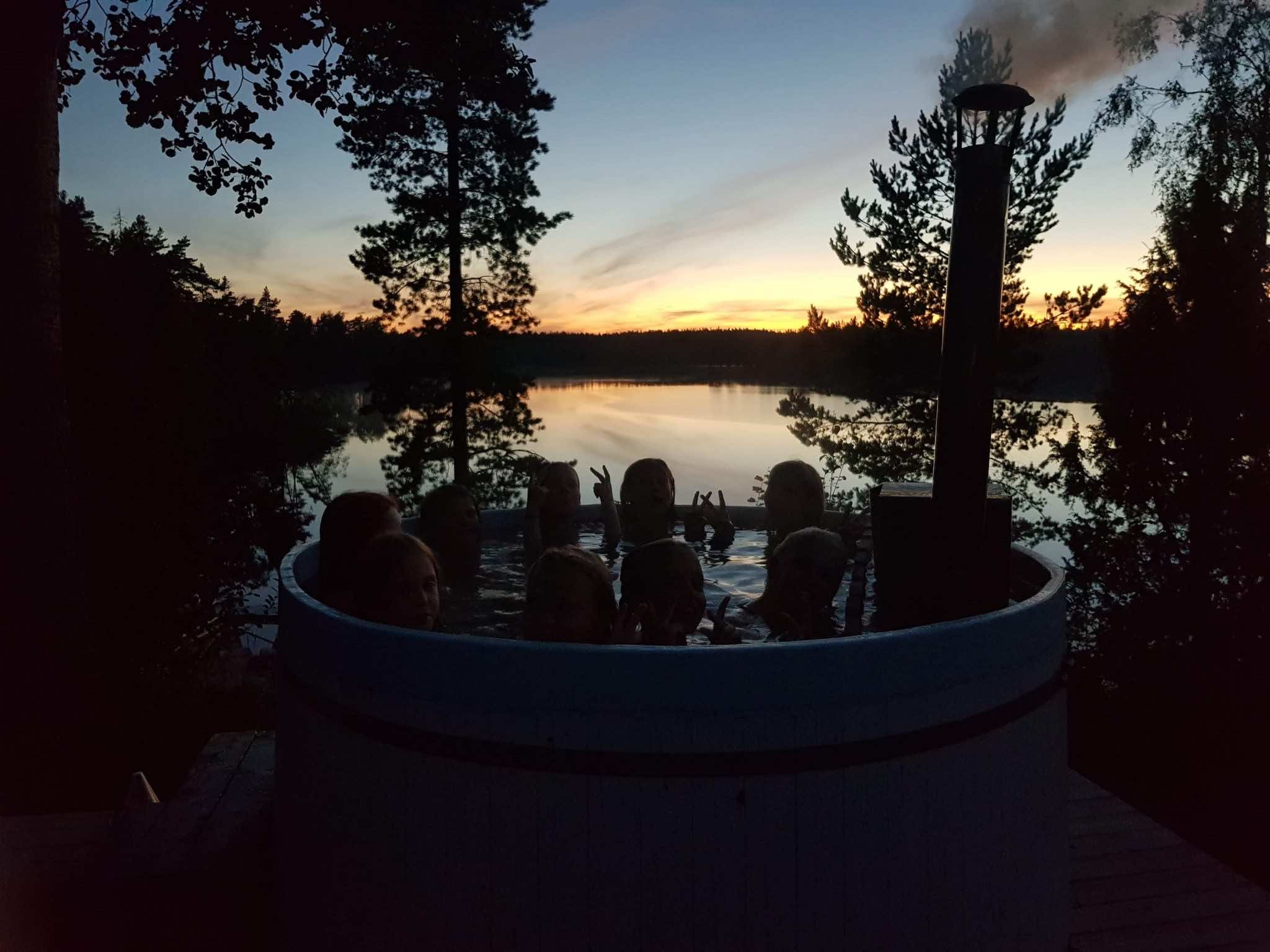 Solnedgång över sjö, ungdomar i en vedeldad badtunna